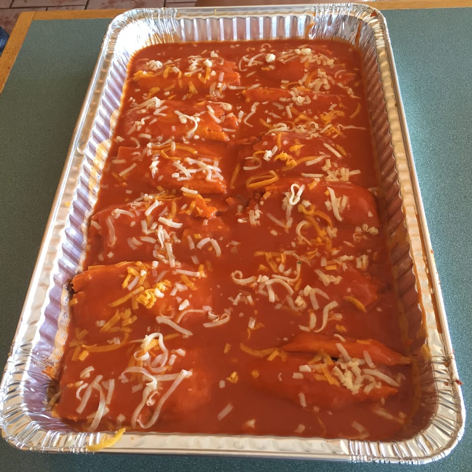 A foil pan filled with enchiladas covered in red sauce and shredded white and yellow cheese.