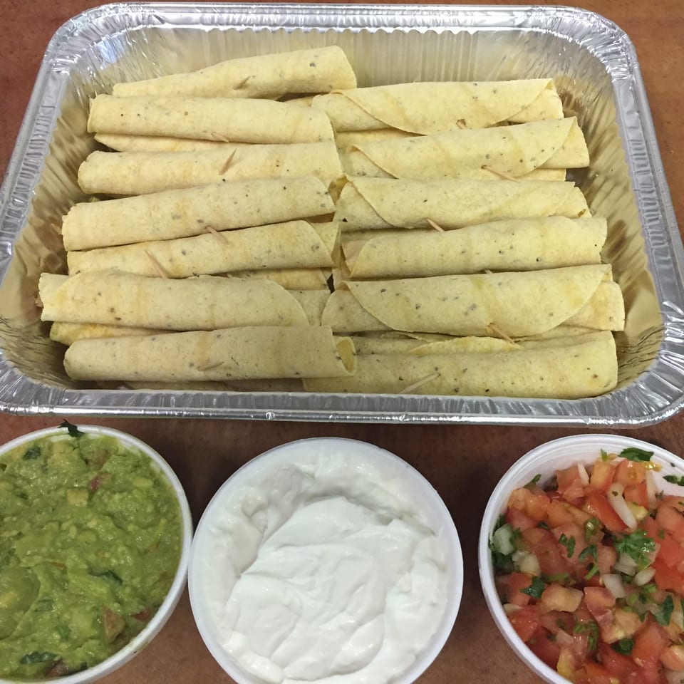 A catering tray of taquitos with sides of guacamole, sour cream, and pico de gallo.